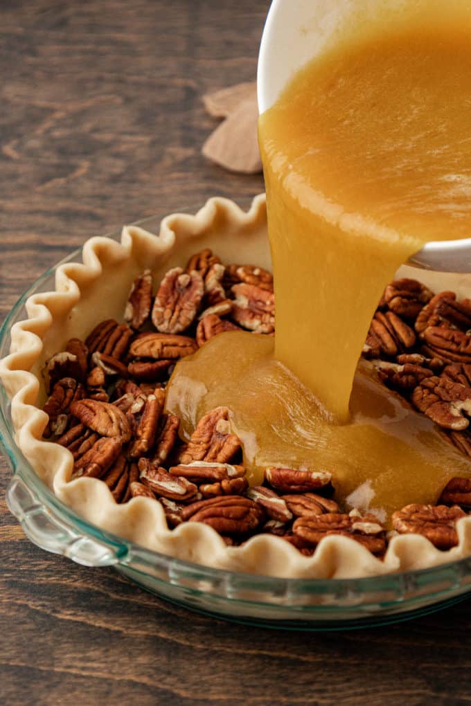 a glass pie dish sitting on a wood surface with pecans in it, and the pecan pie filling mixture being poured over the top of it