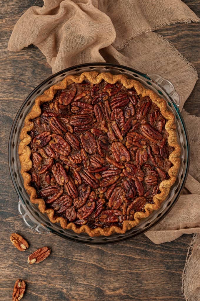 a freshly baked pecan pie in a glass baking dish sitting on a wood surface with a tan towel and whole pecans around it