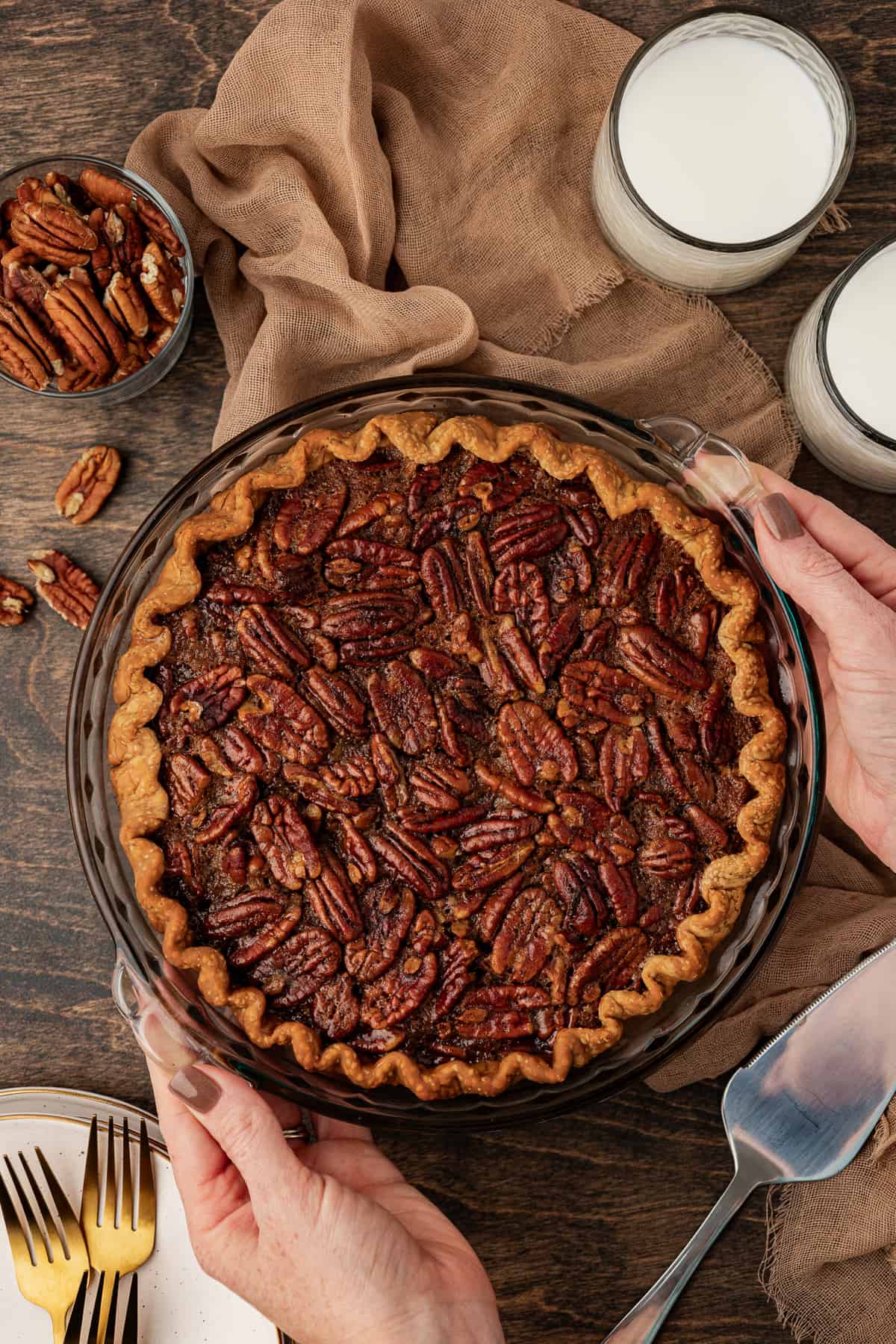 a whole pecan pie being placed on a wood surface with two hands, on top of a tan towel and surrounded by two glasses of milk, a small dish of pecans, more pecans scattered around, a stack of plates and forks and a pie serving spatula
