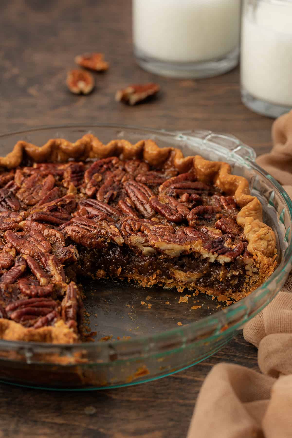 pecan pie with a large slice missing out in a glass pie dish sitting on a wood surface surrounded by pecans and a tan towel with two glasses of milk in the background