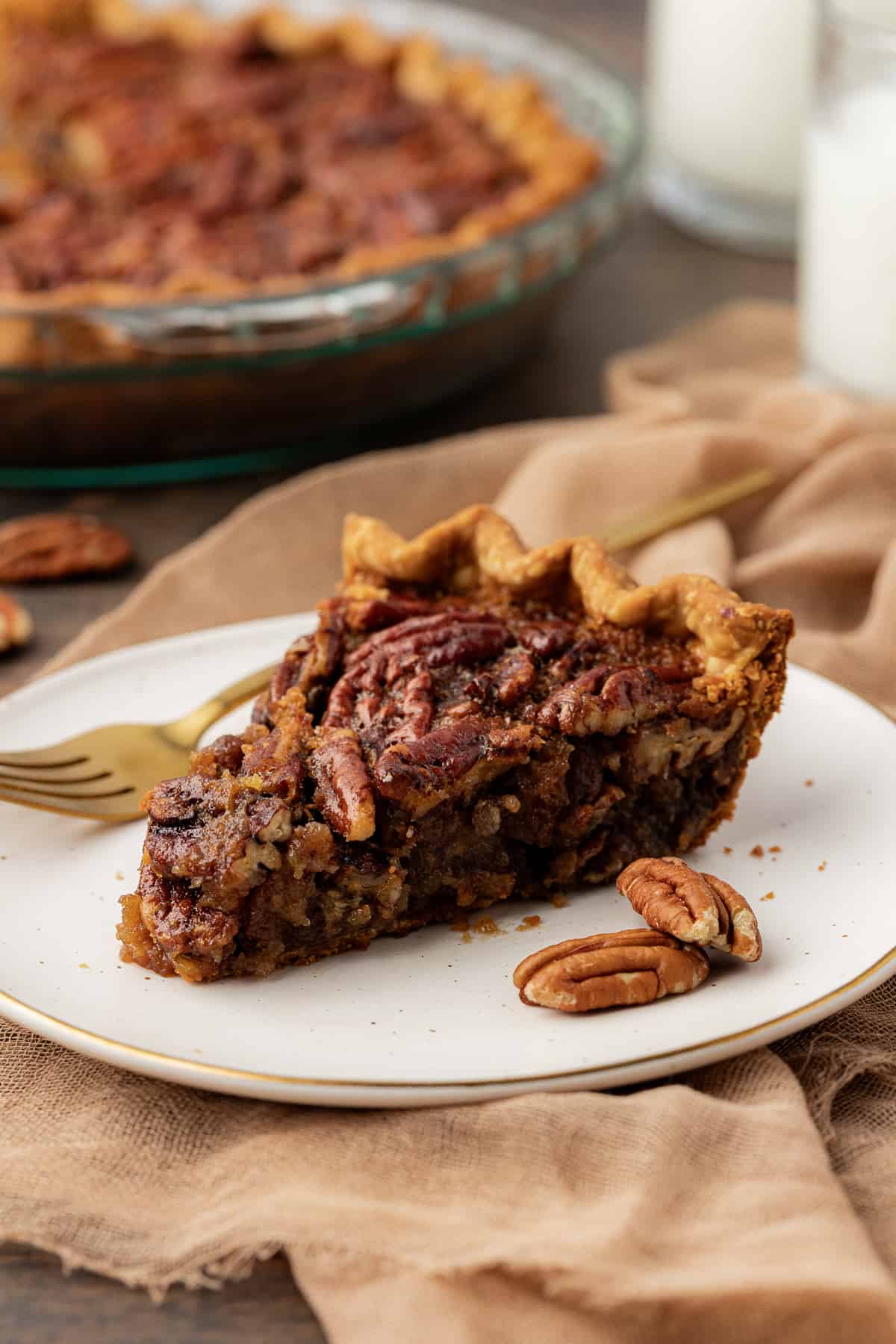 a slice of pecan pie on a white plate with a fork and whole pecans on either side of it, sitting on top of a tan towel on a wood surface, with the glass pie dish full of pie in the background and more pecans scattered around