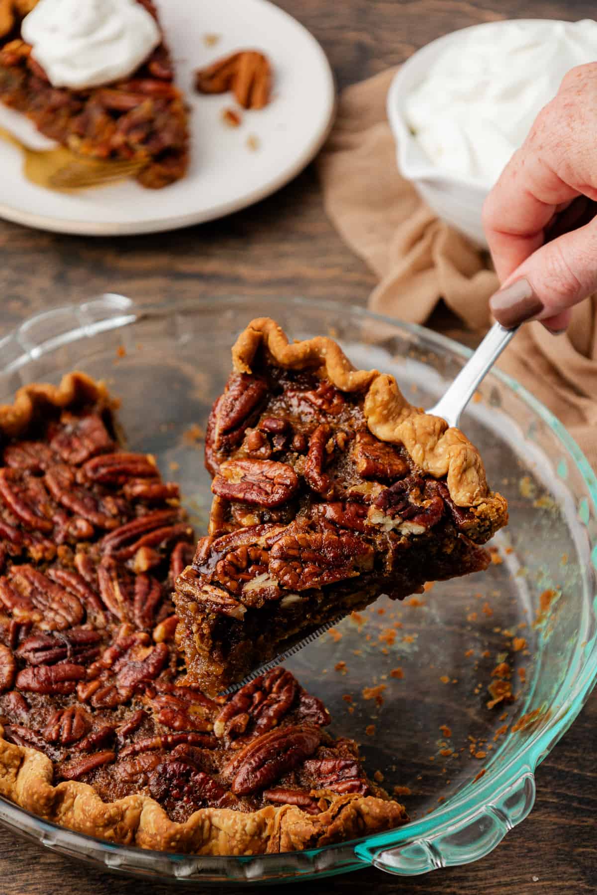a piece of pecan pie being lifted out of a half empty glass pie dish that is sitting on a wood surface beside a tan towel, a plate with a slice of pie topped with whipped cream on it, and a dish of whipped cream. 