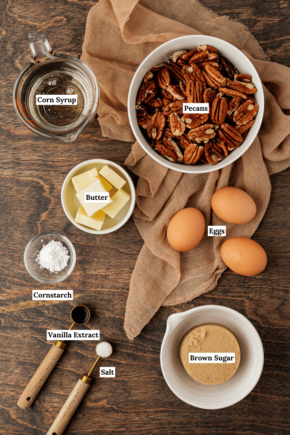 ingredients for pecan pie arranged on a wood surface with a tan towel including corn syrup, pecans, butter, eggs, corn starch, vanilla extract, salt and brown sugar