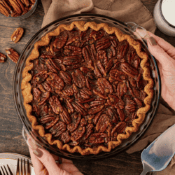 pecan pie in a glass pie dish on a wood surface with a tan towel, pecans, a serving spatula, plates and forks surrounding it