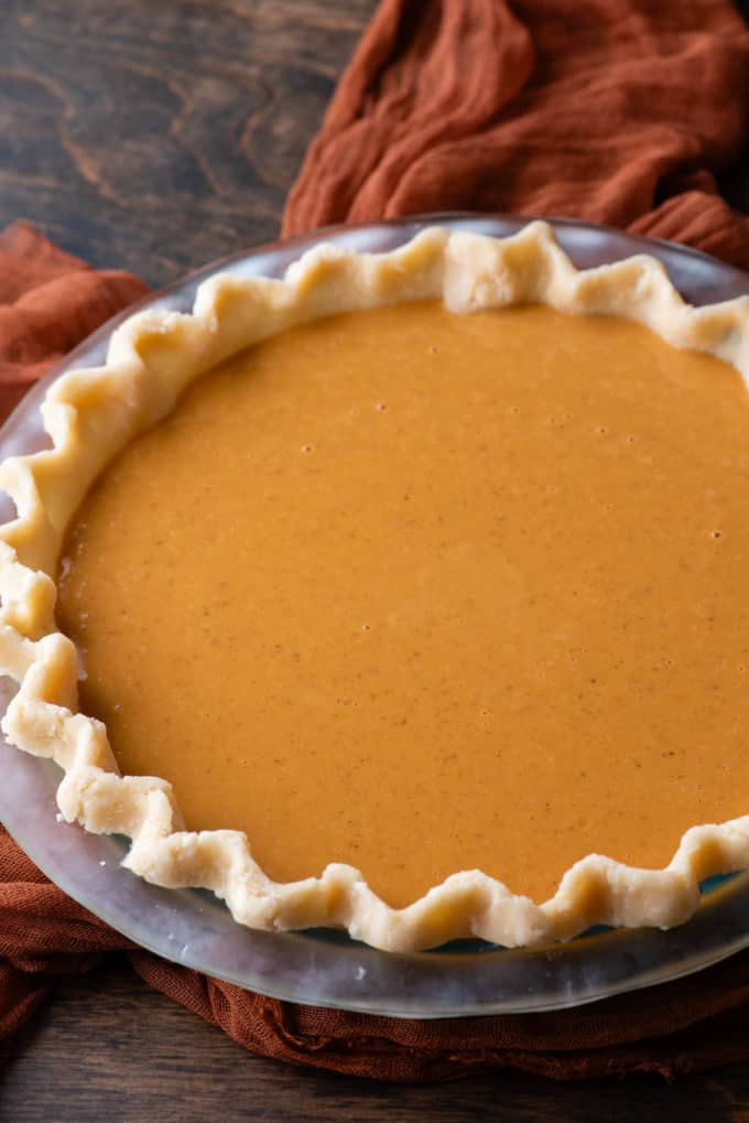 an uncooked pumpin pie in a glass pie dish sitting on a dark orange cloth on a wood surface