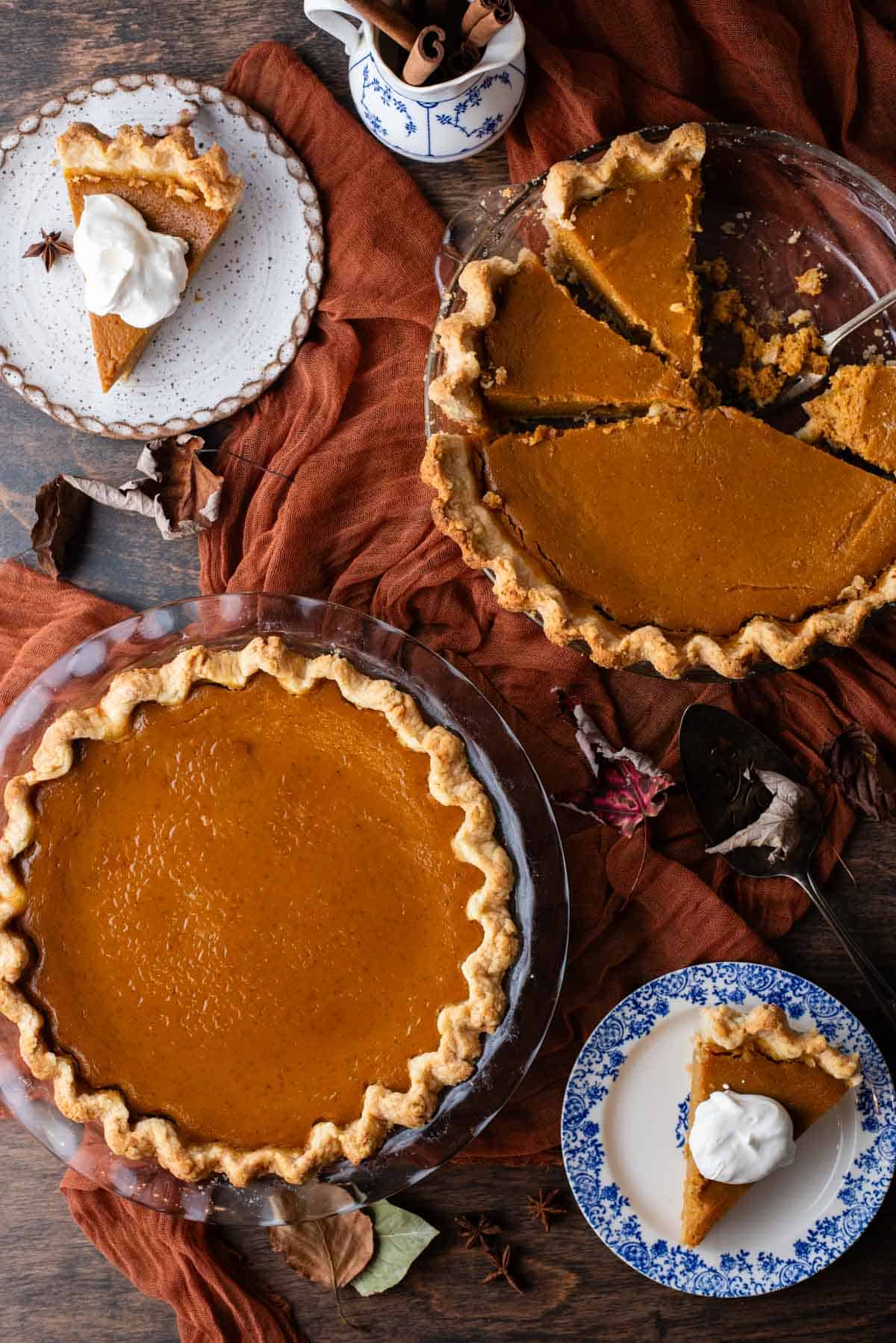 an overhead view of a tablescape with two pumpkin pies, one whole and one sliced, sitting on top of a dark orange cloth on a wood surface and surrounded by two small plates with pumpkin pie slices topped with whipped cream, fall leaves and a dish of cinnamon sticks