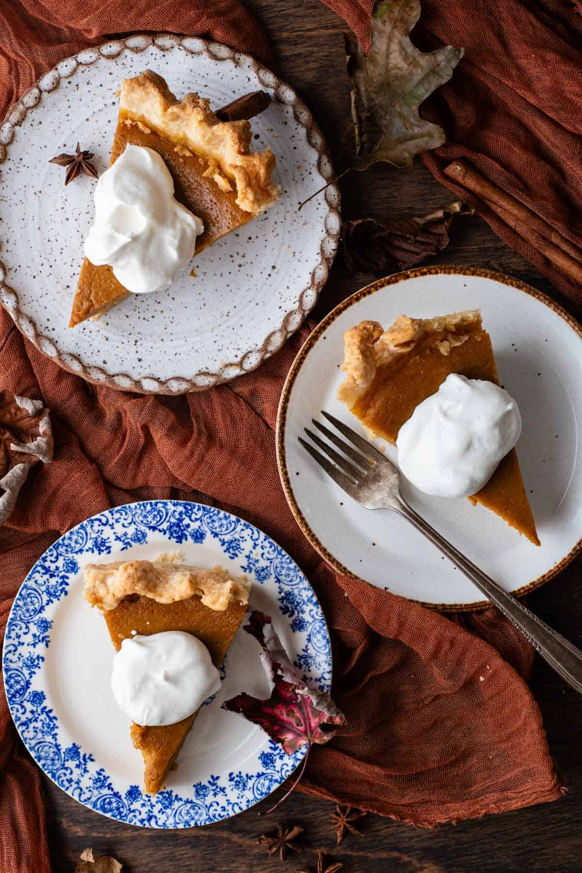 three small plates sitting on a wood surface with a dark orange cloth and fall leaves scattered around, with slices of pumpkin pie on them topped with a dollop of whipped cream, and a fork on the middle plate
