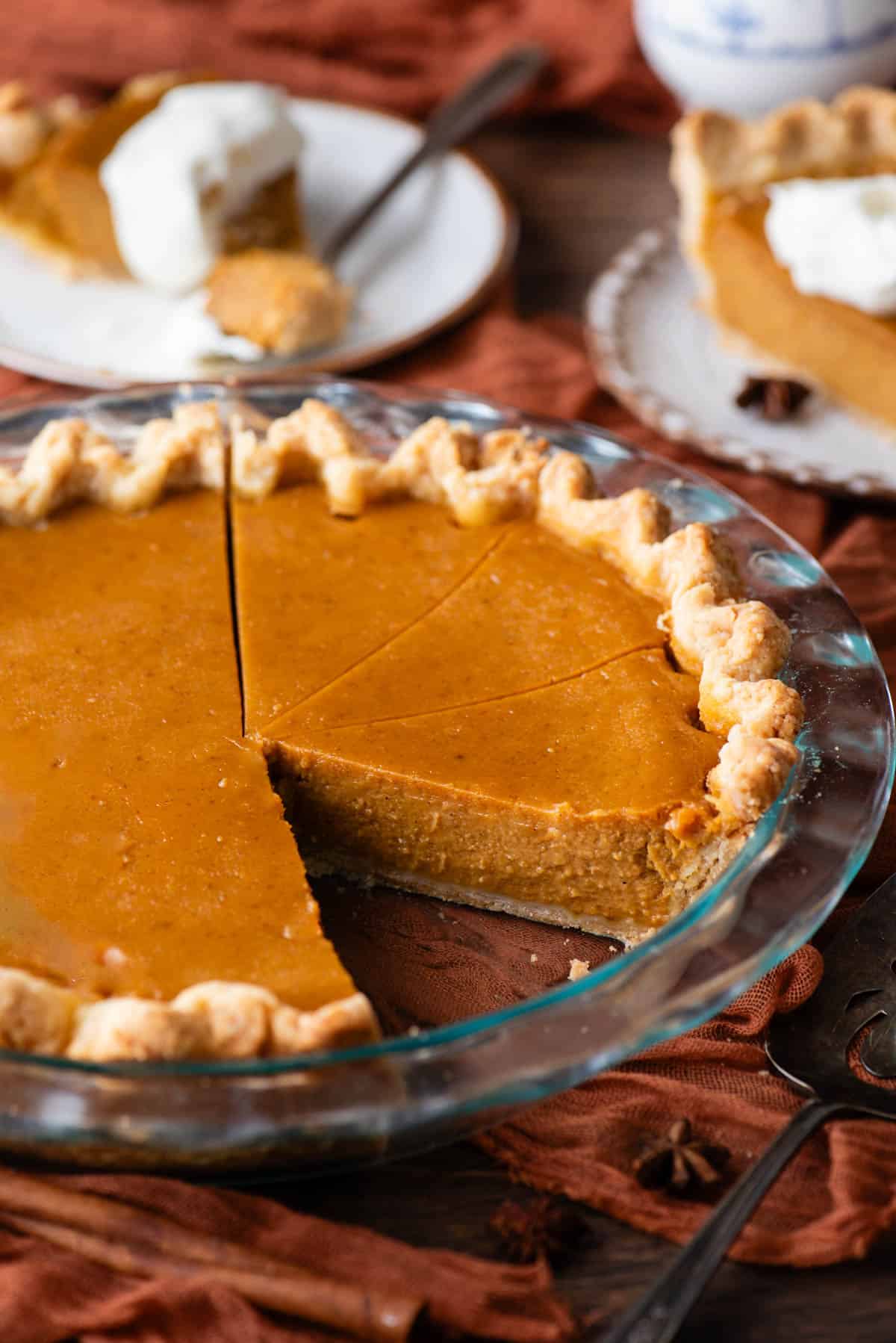 pumpkin pie in a glass pie dish on a wood surface with a dark orange cloth under it, sliced with one slice missing and two plates in the background with pumpkin pie on them