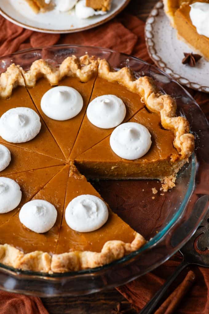 a pumpkin pie in a glass pie dish with dollops of whipped cream on top, with one slice missing, sitting on top of a dark orange cloth on a wood surface with two plates with pumpkin pie on them beside it