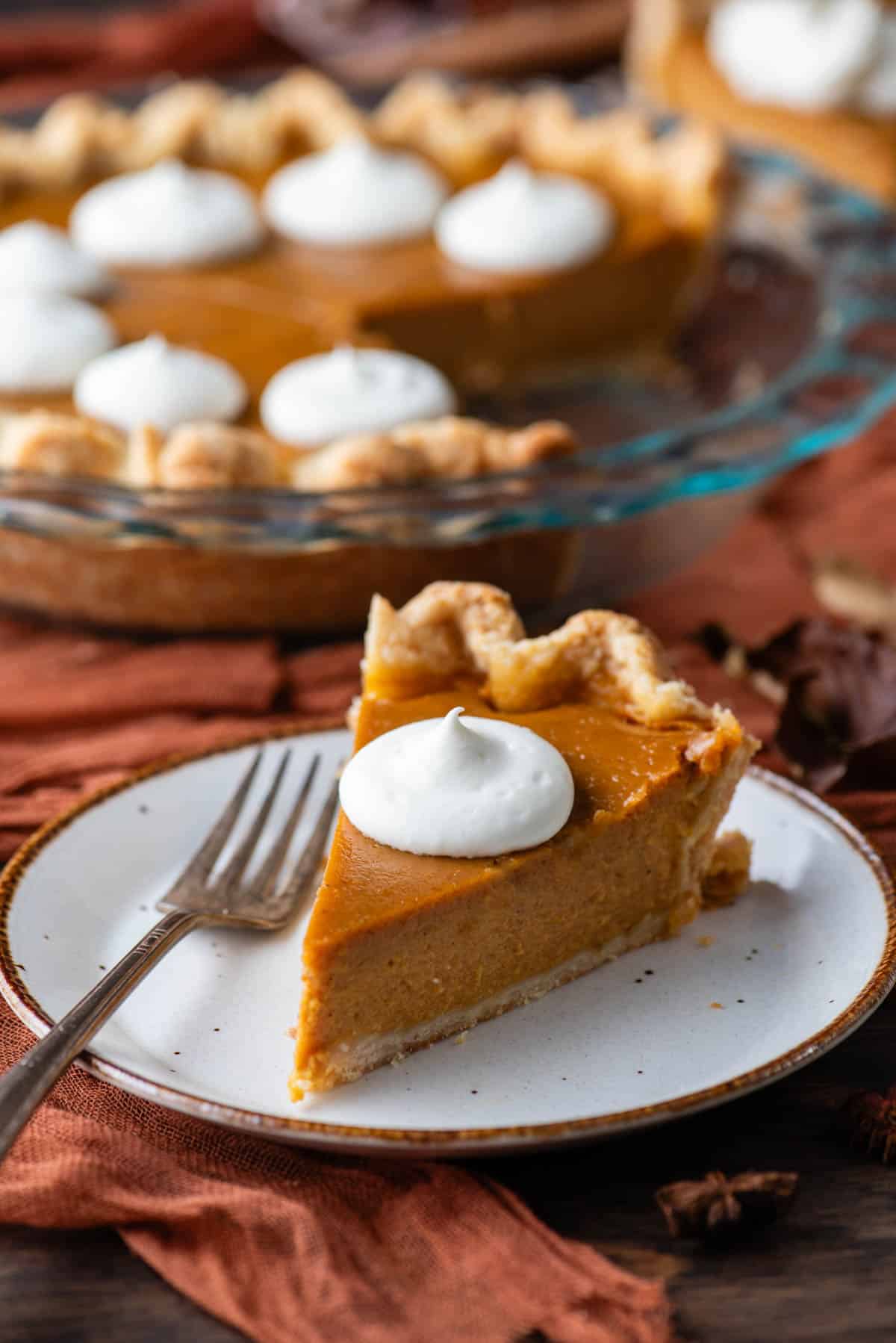 a slice of pumpkin pie on a plate topped with whipped cream with a fork sitting beside it on the plate, with the rest of the pie behind it in a glass pie dish, all sitting on a wood surface with a dark orange cloth
