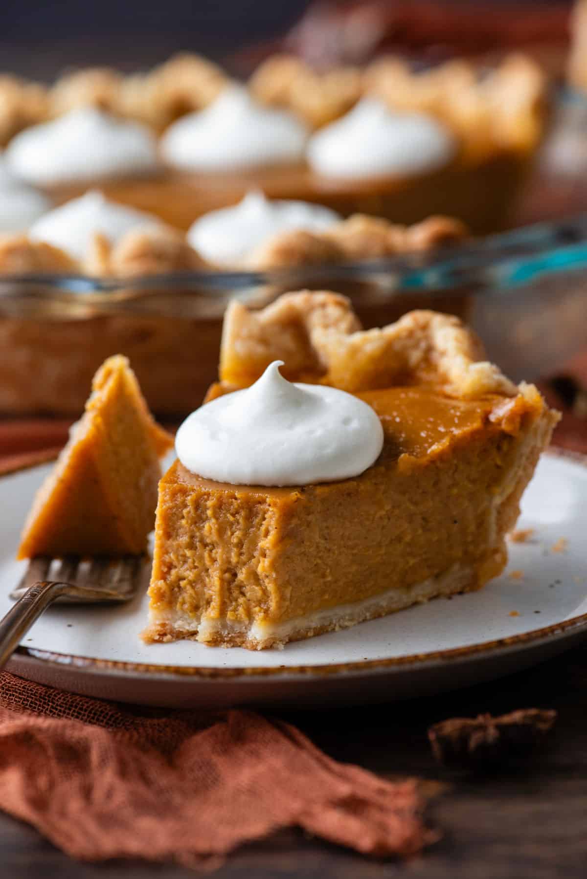 a slice of pumpkin pie on a white plate topped with a dollop of whipped cream, with a fork that has taken one bite out laying on the plate beside it, and the pie dish with the rest of the pie in the background