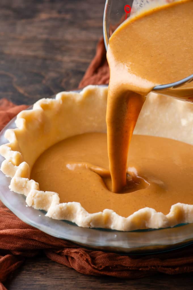 pumpkin pie filling being poured into a glass pie dish with pie crust in it, which is sitting on top of a dark orange cloth on a wood surface