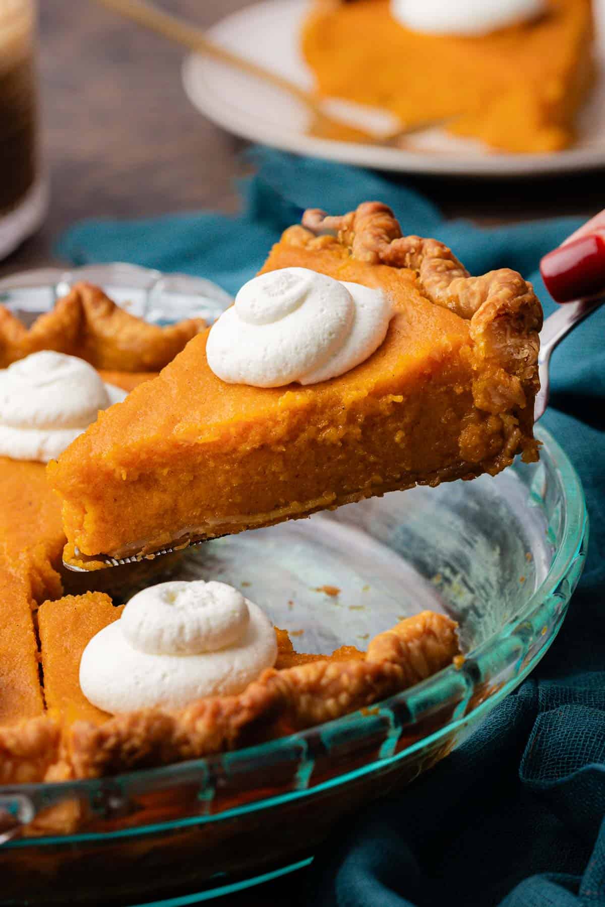 a slice of sweet potato pie being lifted out of a glass pie dish with more slices of pie in it, all topped with dollops of whipped cream