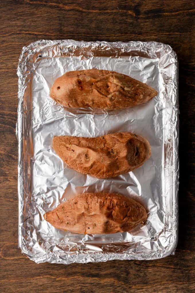 three roasted sweet potatoes on a sheet pan covered in aluminum foil sitting on a wood surface