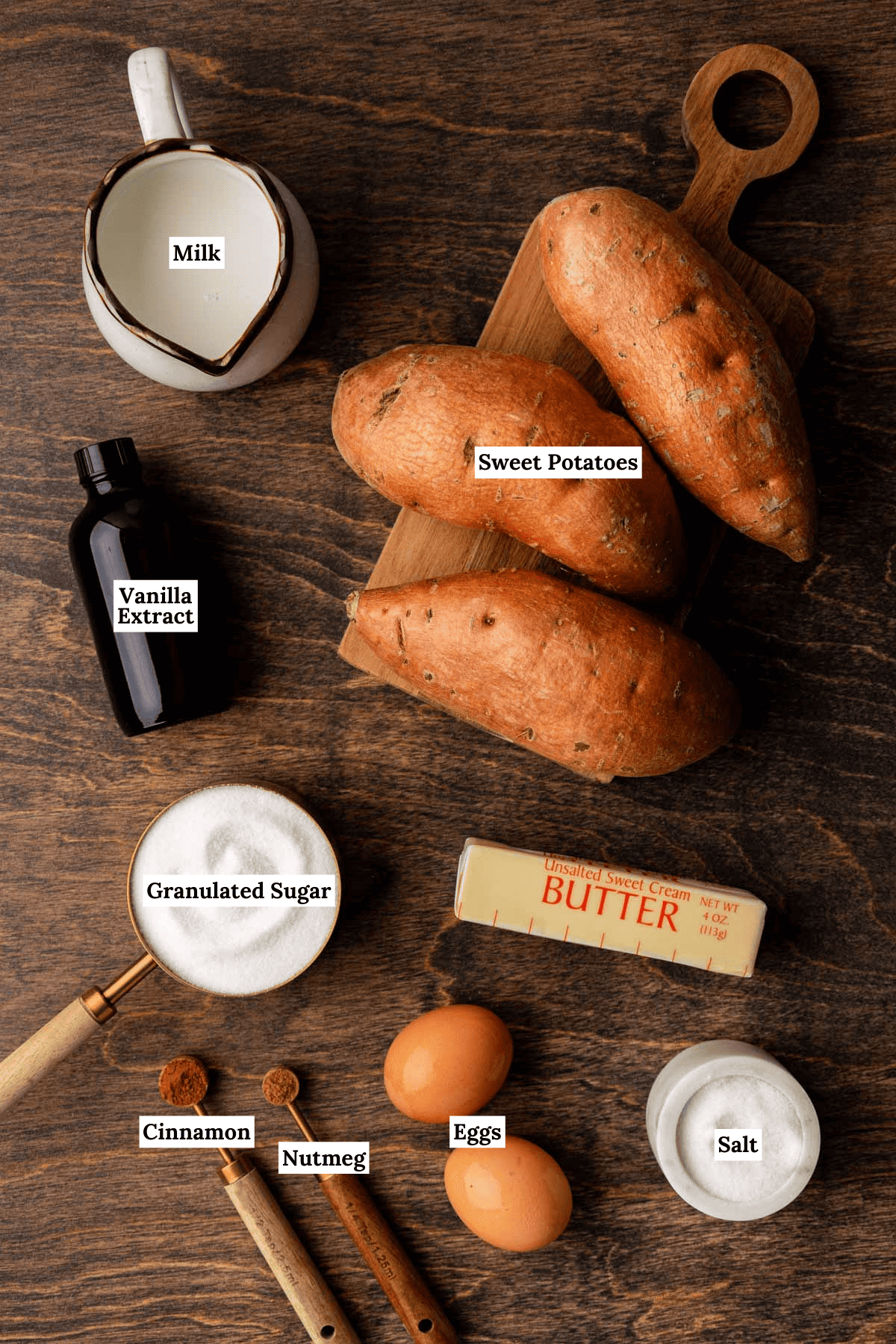 ingredients for sweet potato pie arranged on a wood surface including whole sweet potatoes, milk, vanilla extract, granulated sugar, butter, eggs, salt, nutmeg and cinnamon