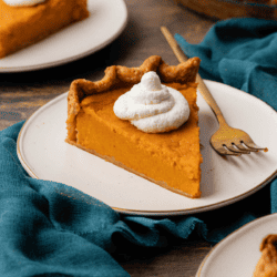 slices of sweet potato pie topped with dollops of whipped cream on small white places on a dark teal towel on a wood surface with a fork laying beside the pie on the middle plate