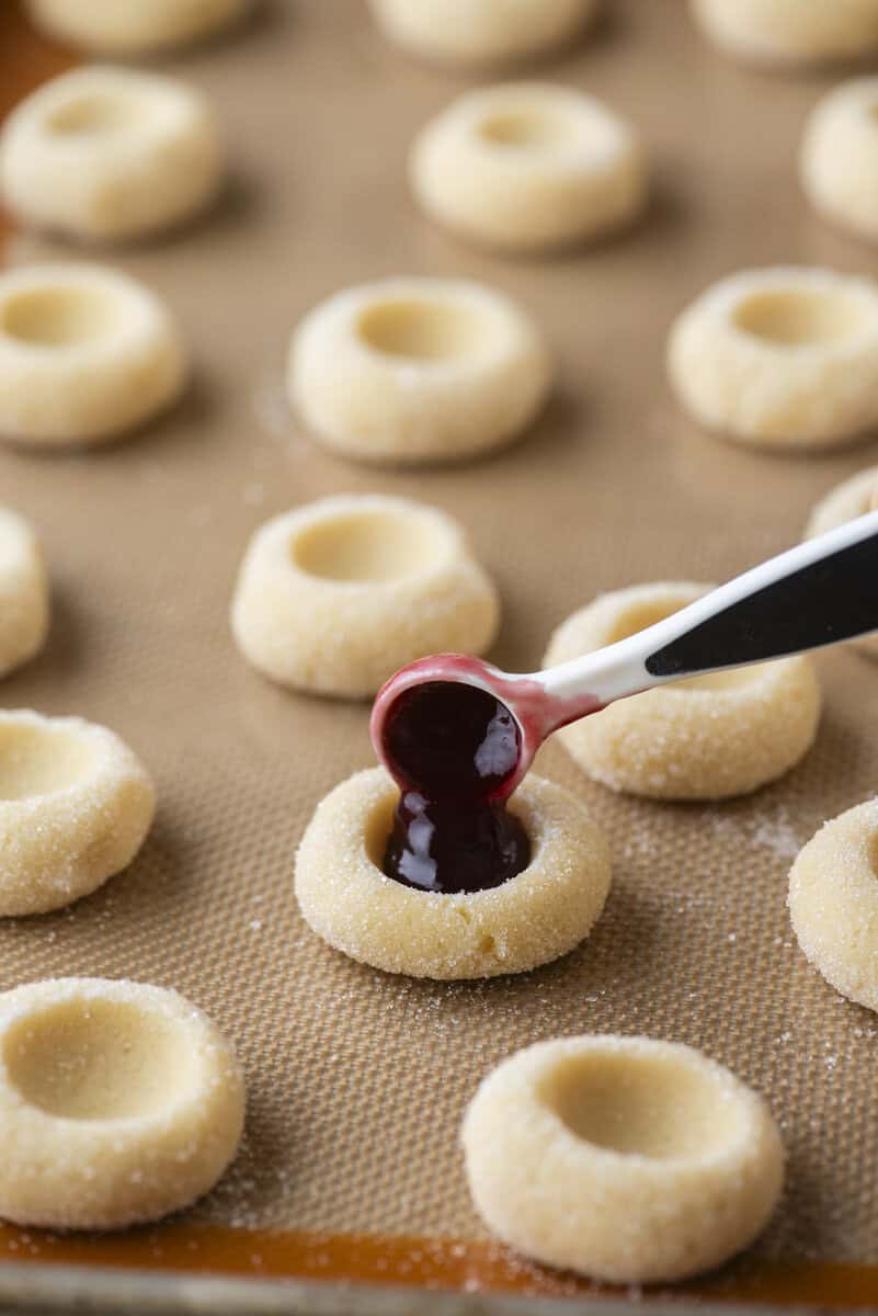 A hand uses a small spoon to fill delicious thumbprint cookies with red jam. Several unfilled thumbprint cookies with indents rest on a baking sheet lined with a silicone mat.