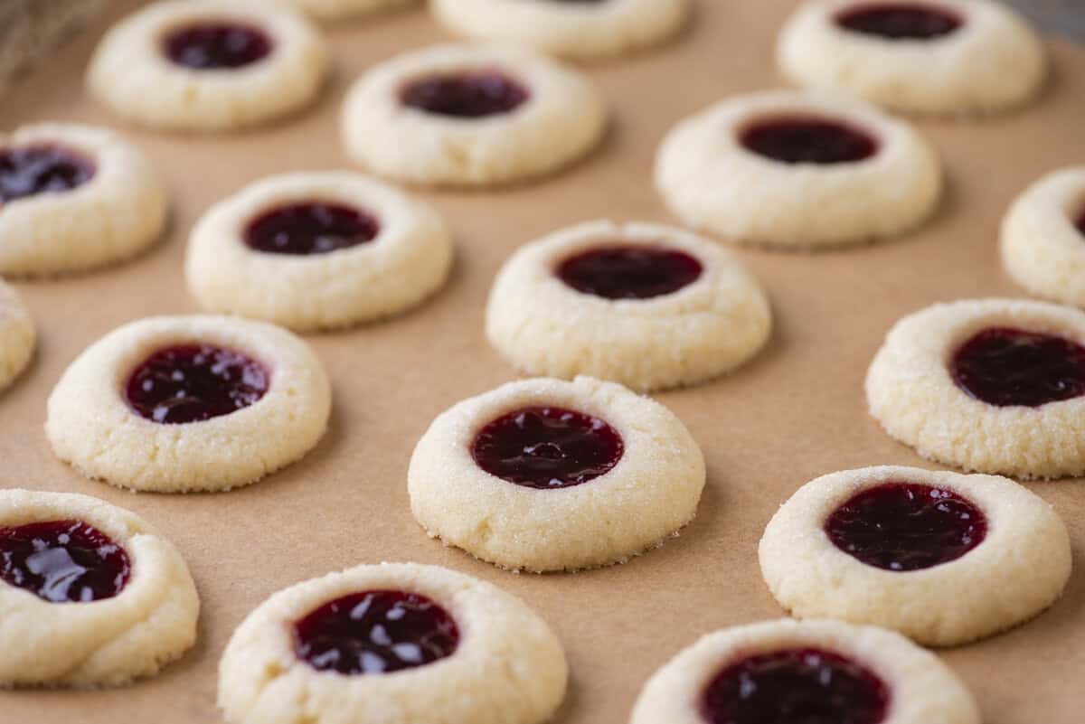 Rows of round thumbprint cookies with red jam centers are arranged on a sheet of brown parchment paper. The crumbly, sugar-coated thumbprint cookies are evenly spaced, showing off their classic holiday charm.