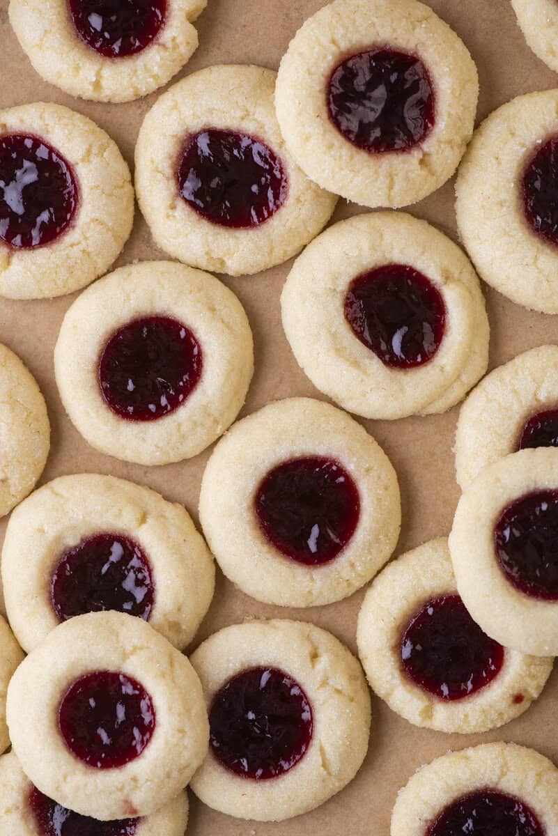A close-up of several thumbprint cookies with a red jam filling in the center, arranged on brown parchment paper. The thumbprint cookies have a soft, crumbly texture.