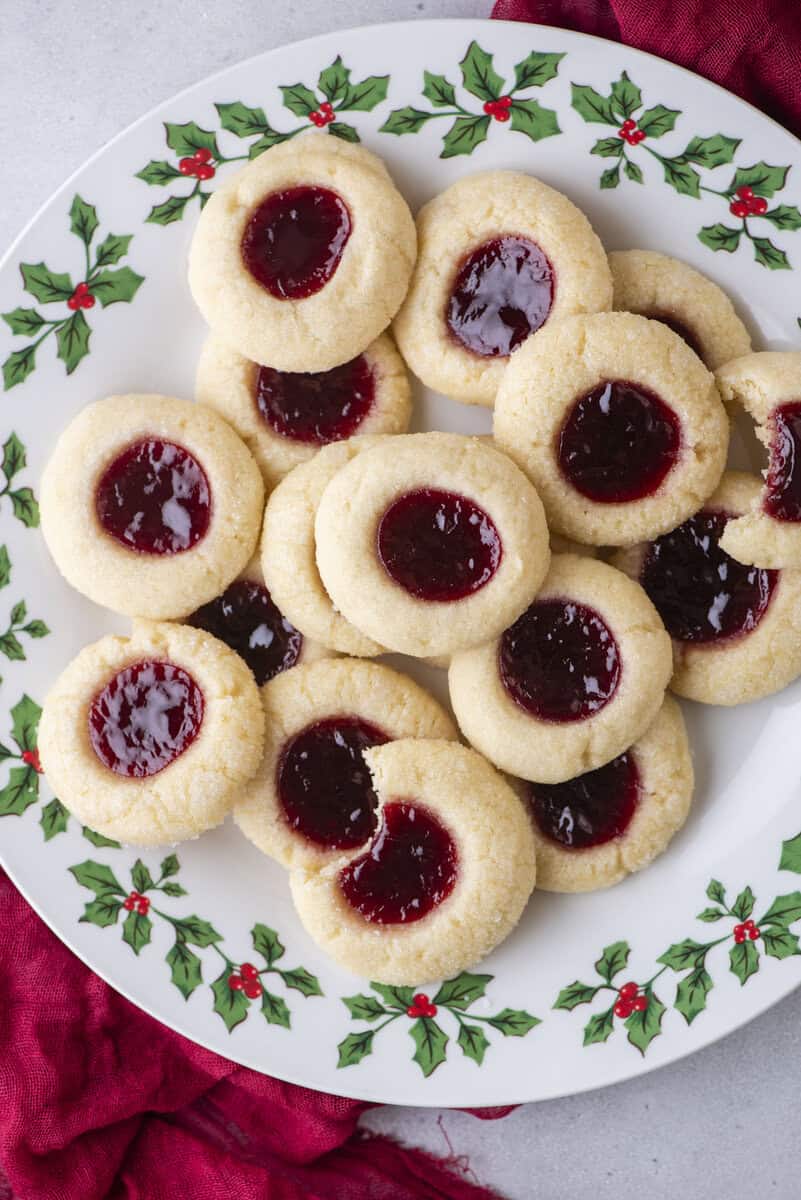 A festive plate decorated with holly holds several classic thumbprint cookies filled with red jam, arranged in a pile on a light surface with a red cloth partially visible.