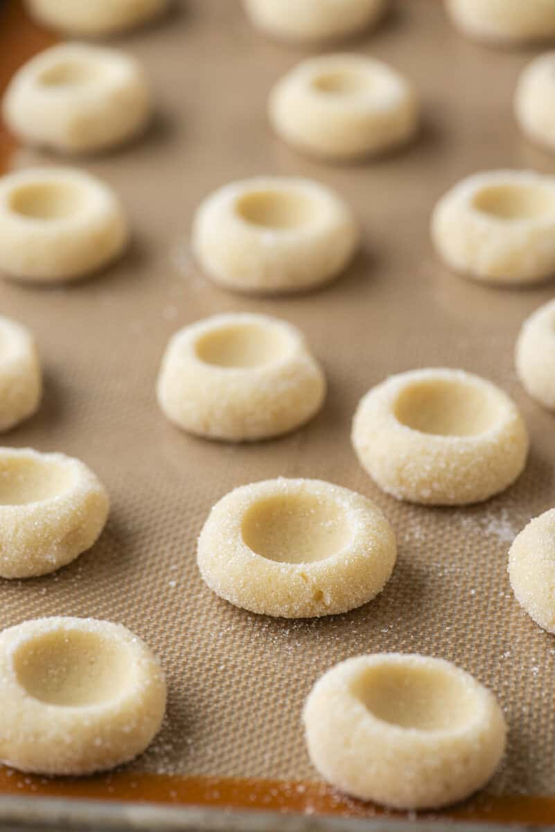 Rows of unbaked thumbprint cookies with indented centers are arranged on a baking sheet lined with a silicone mat. The thumbprint cookies are coated lightly with sugar and spaced evenly apart.