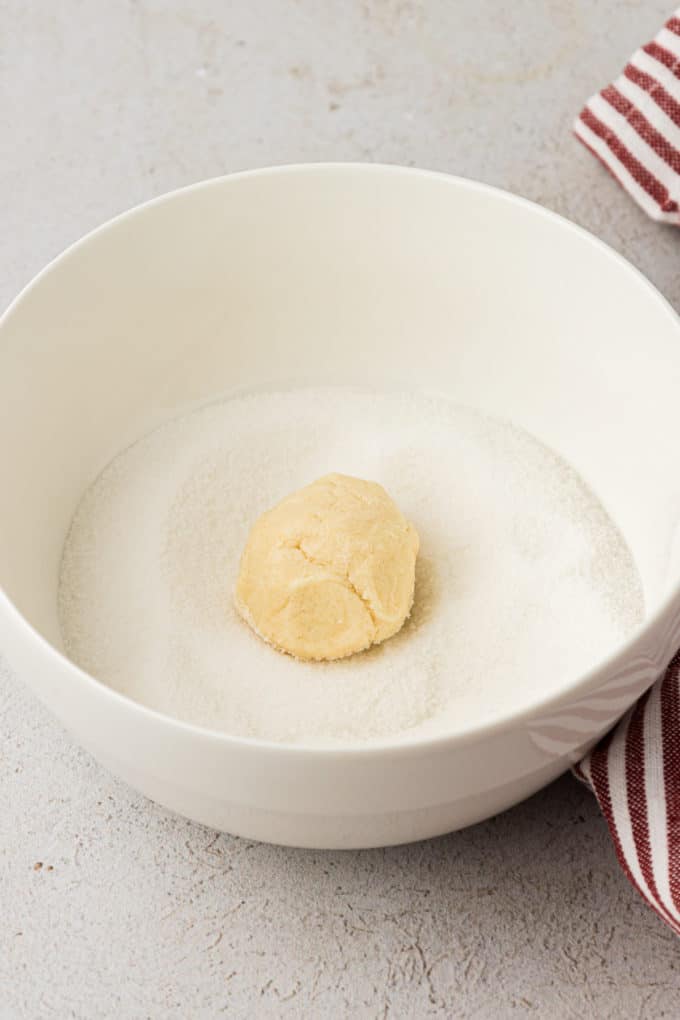 thumbprint cookie dough formed into a ball laying in a white bowl of sugar on a countertop beside a red and white striped towel
