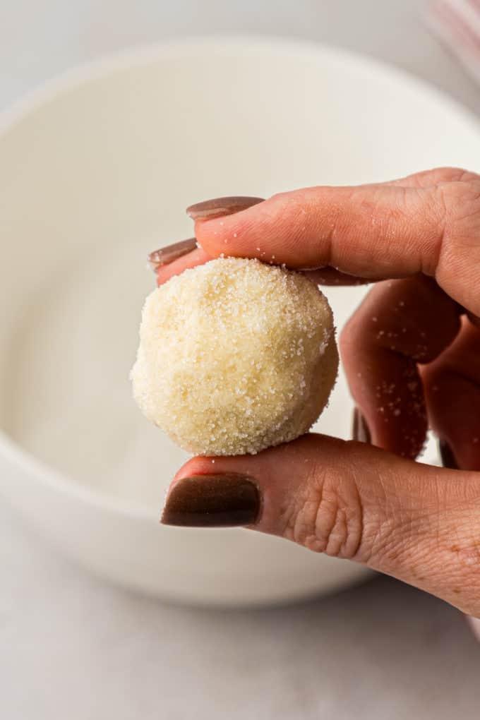 a hand holding a ball of thumbprint cookie dough that has been rolled in sugar over top of the white bowl of sugar in the background