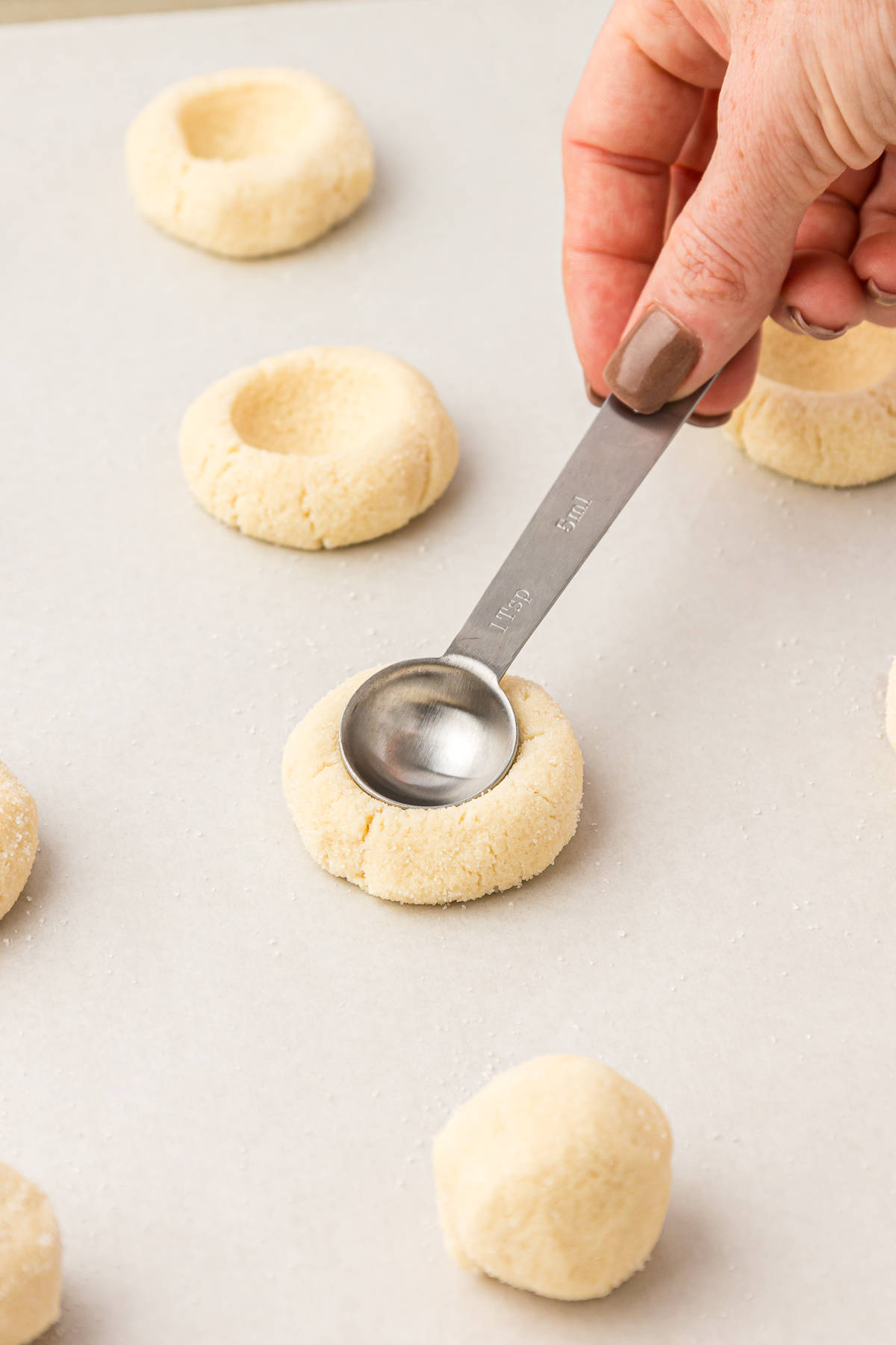thumbprint cookie dough balls on a baking sheet lined with parchment paper with indentations being pressed in by the back of a spoon