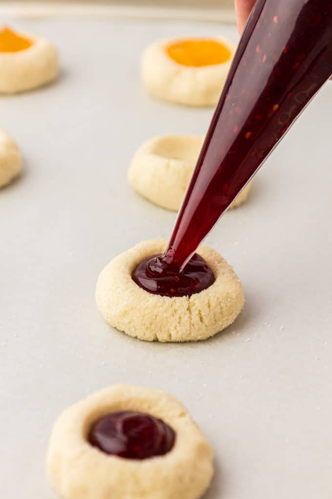 thumbprint cookies on a baking sheet lined with parchment paper being filled with raspberry jam