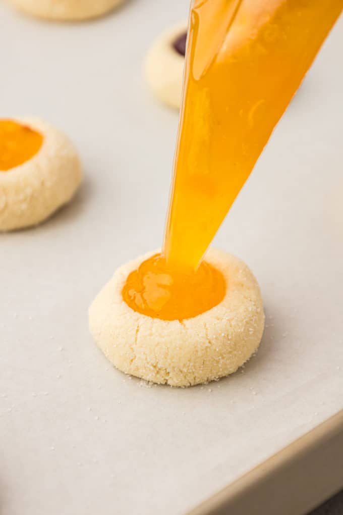 thumbprint cookies on a baking sheet lined with parchment paper being filled with apricot jam