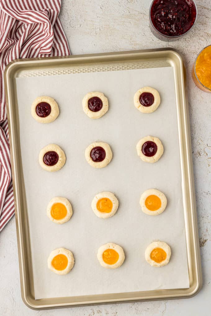 thumbprint cookies filled with raspberry and apricot jam on a baking sheet lined with parchment paper sitting on a countertop by a red and white striped towel and jars of raspberry and apricot jam