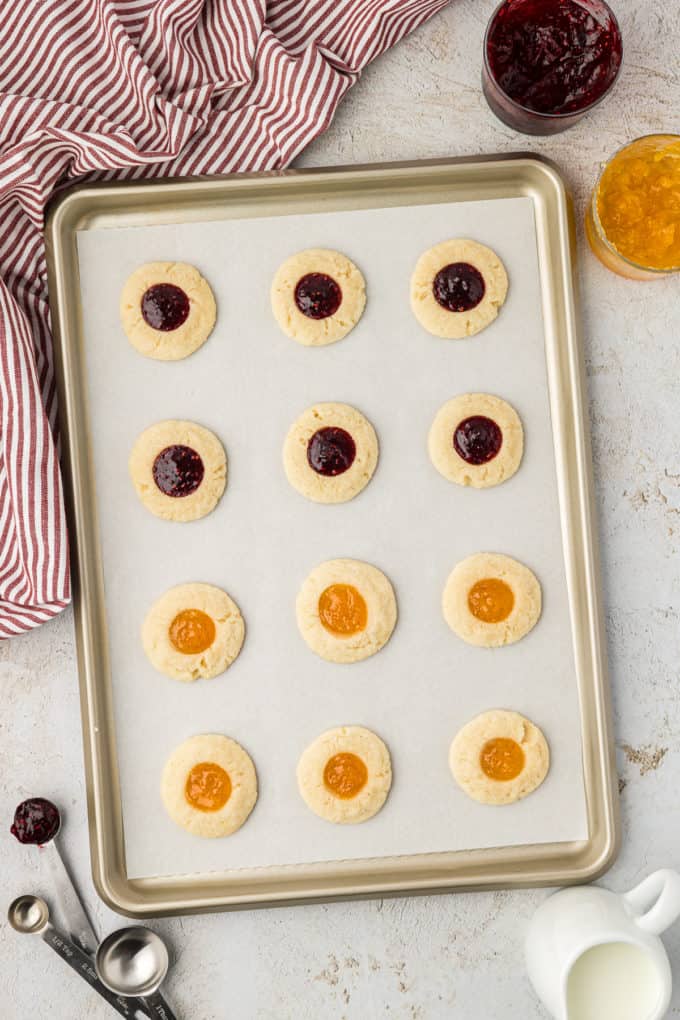 freshly baked thumbprint cookies filled with raspberry and apricot jam on a baking sheet lined with parchment paper sitting on a countertop by a red and white striped towel and jars of raspberry and apricot jam
