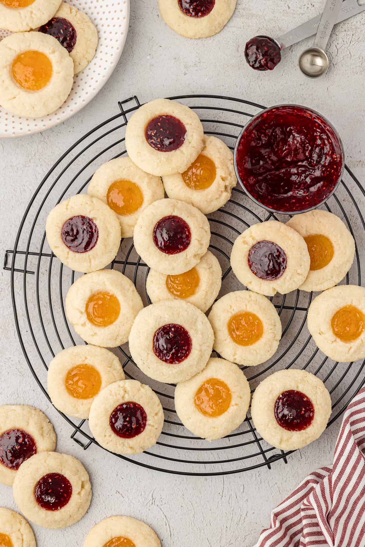 apricot and raspberry jam thumbprint cookies scattered on a wire rack beside a jar of raspberry jam, with more cookies scattered around, a plate of cookies, measuring spoons and a red and white striped towel