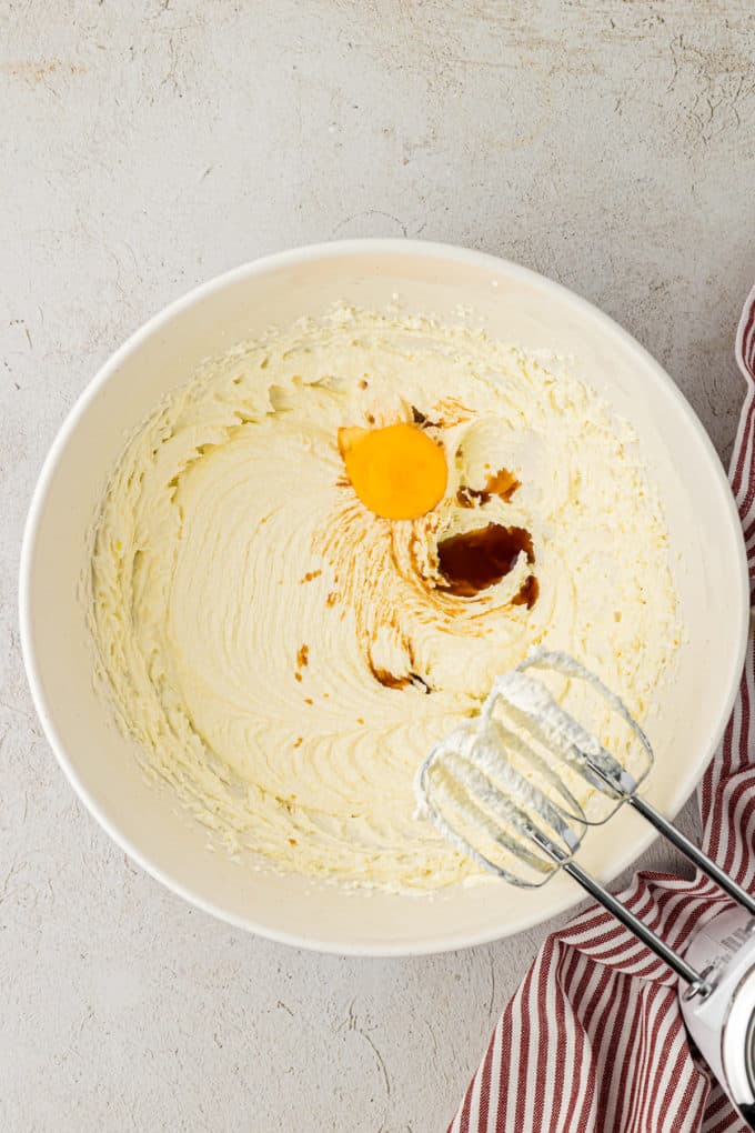 butter, sugar egg yolk and vanilla extra in a white bowl on a countertop beside a red and white striped towel with an electric mixer leaning on the side of the bowl