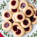 A plate decorated with holly displays a festive pile of thumbprint cookies filled with red jam. One thumbprint cookie has a bite taken out, revealing its soft, tender texture inside.