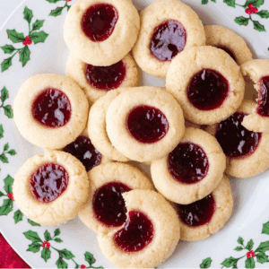A plate decorated with holly displays a festive pile of thumbprint cookies filled with red jam. One thumbprint cookie has a bite taken out, revealing its soft, tender texture inside.