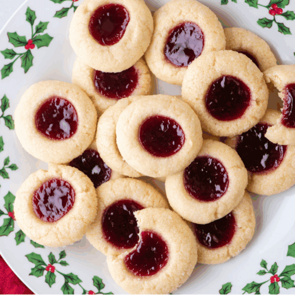 A plate decorated with holly displays a festive pile of thumbprint cookies filled with red jam. One thumbprint cookie has a bite taken out, revealing its soft, tender texture inside.