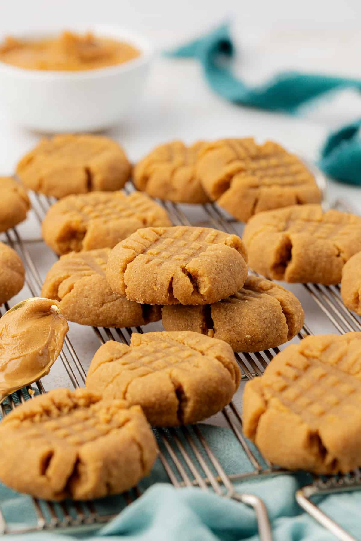 peanut butter cookies piled on a wire rack on top of a light teal towel with a bowl of peanut butter in the background