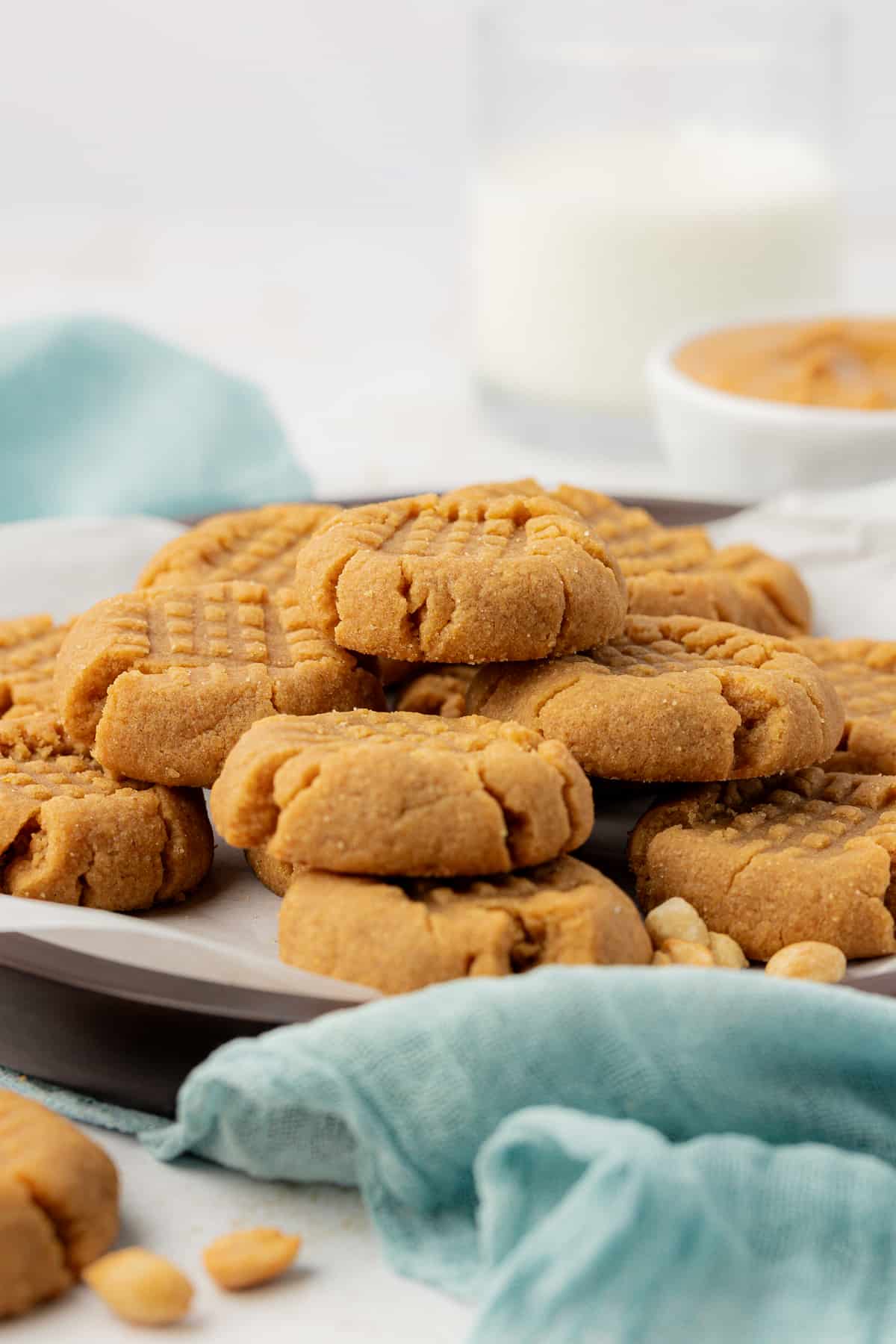 a plate full of 3 ingredient peanut butter cookies with a teal kitchen towel around it and a bowl of peanut butter and a cup of milk in the background