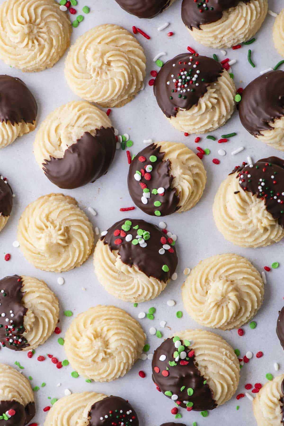 An assortment of butter cookies lined in rows on parchment paper, some plain cookies, some dipped in chocolate and some dipped in chocolate with red, green and white sprinkles, with sprinkles scattered around between the cookies