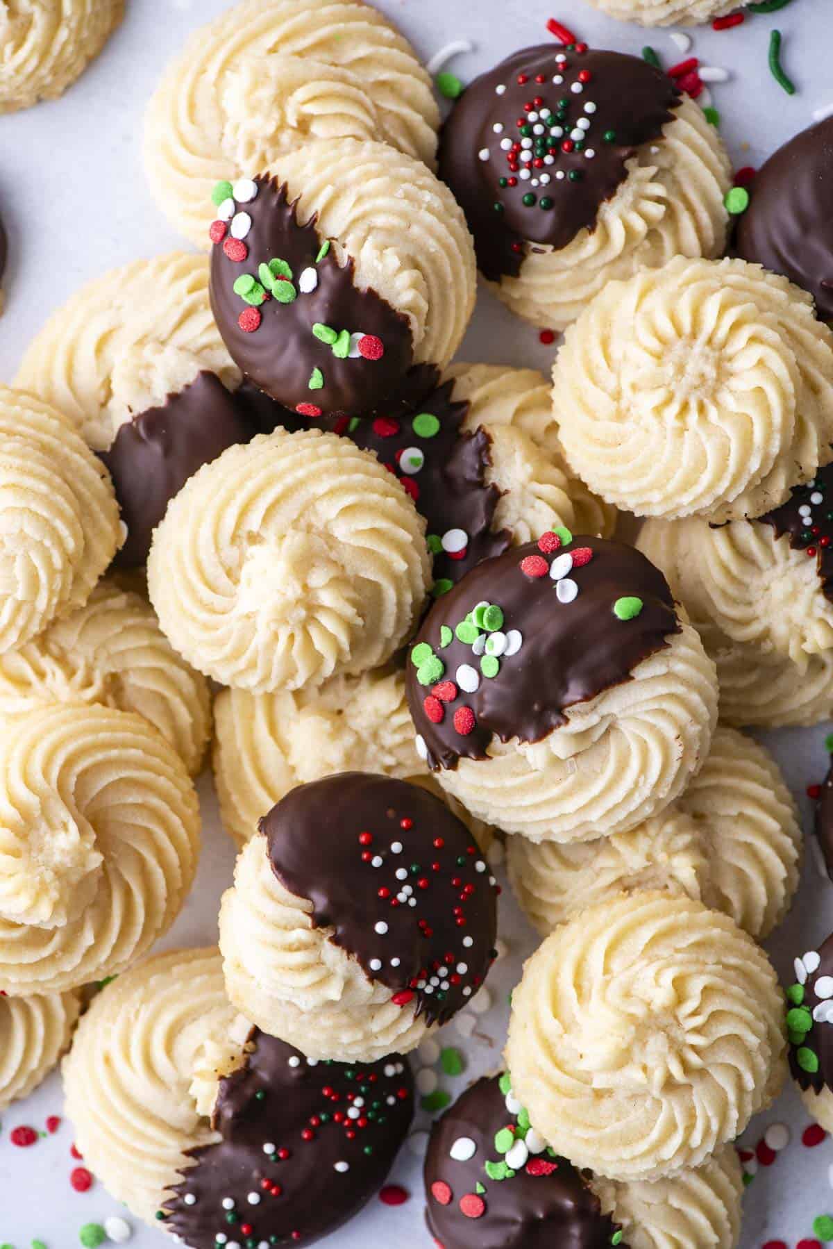 An assortment of butter cookies piled on parchment paper, some plain cookies, some dipped in chocolate and some dipped in chocolate with red, green and white sprinkles