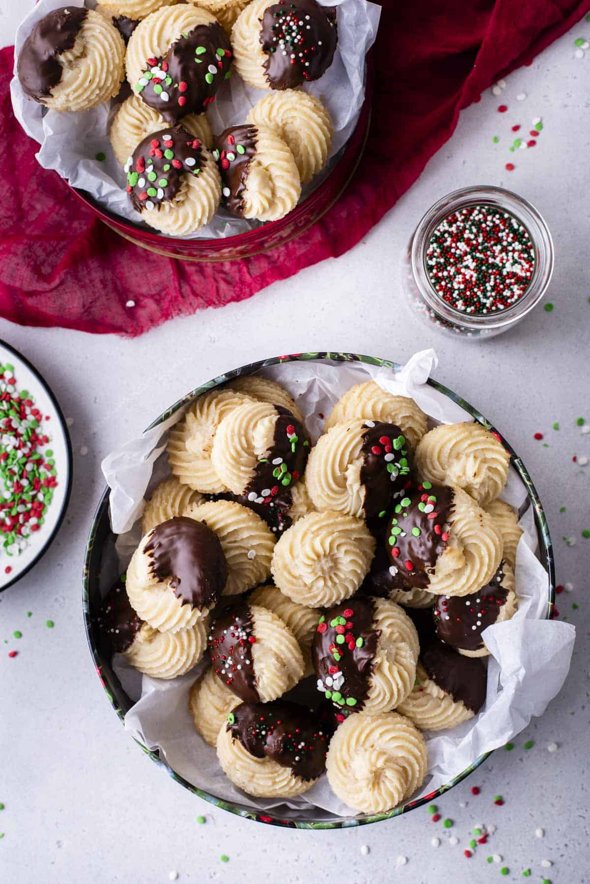 two christmas tins full of butter cookies with a bowl of sprinkles and a small glass dish of sprinkles. Some of the cookies are plain, some dipped in chocolate and some dipped in chocolate with red, green and white sprinkles