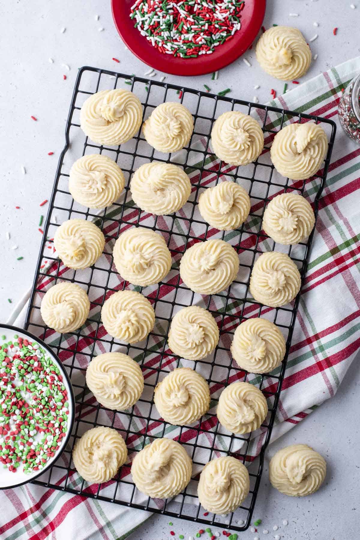 plain butter cookies in rows on a wire rack on top of a red white and green kitchen towel with a bowl of red, white and green sprinkles