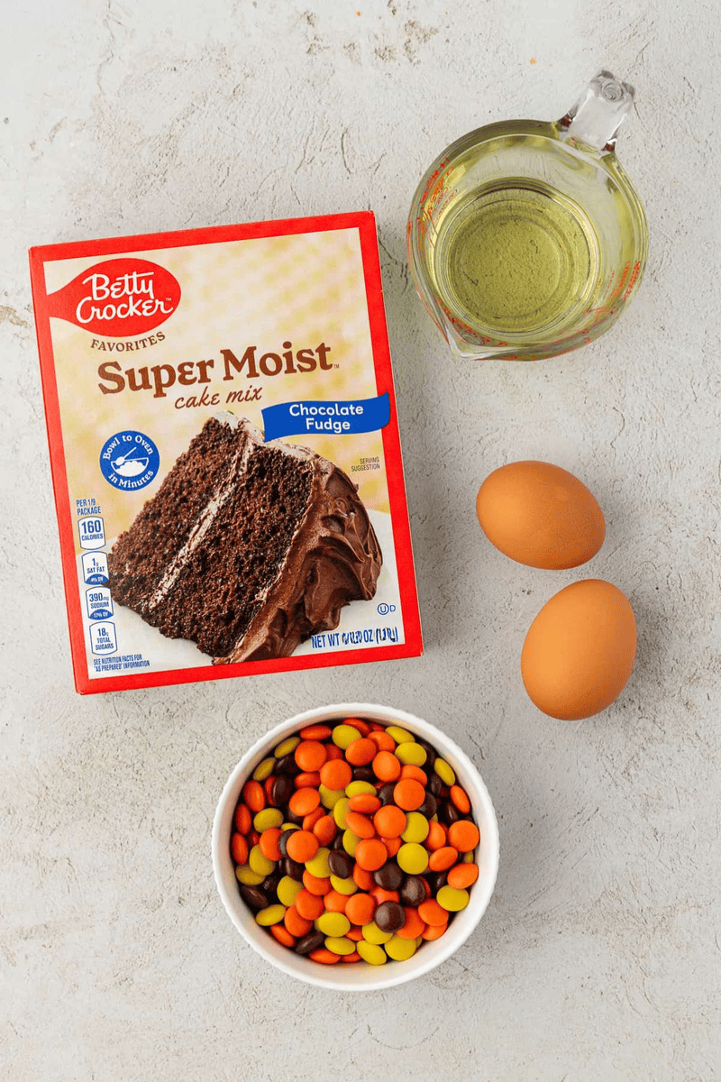 A box of Betty Crocker Super Moist Chocolate Fudge cake mix, a glass measuring cup of oil, two eggs, and a bowl of colorful peanut butter candies are arranged on a light surface—perfect for making delicious cake mix cookies.