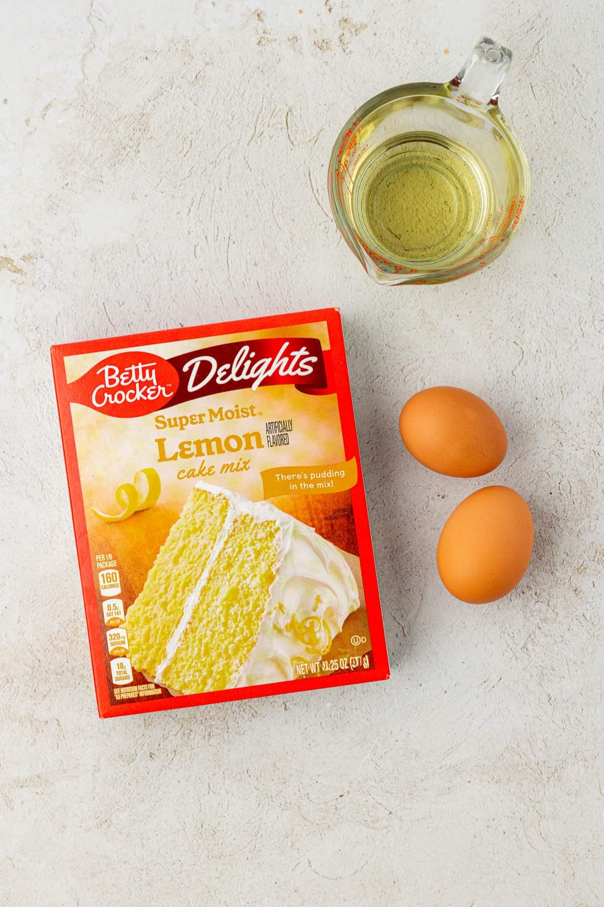 A box of Betty Crocker Delights Super Moist Lemon cake mix, a glass measuring cup with oil, and two brown eggs are arranged on a light-colored surface—perfect for whipping up easy cake mix cookies.
