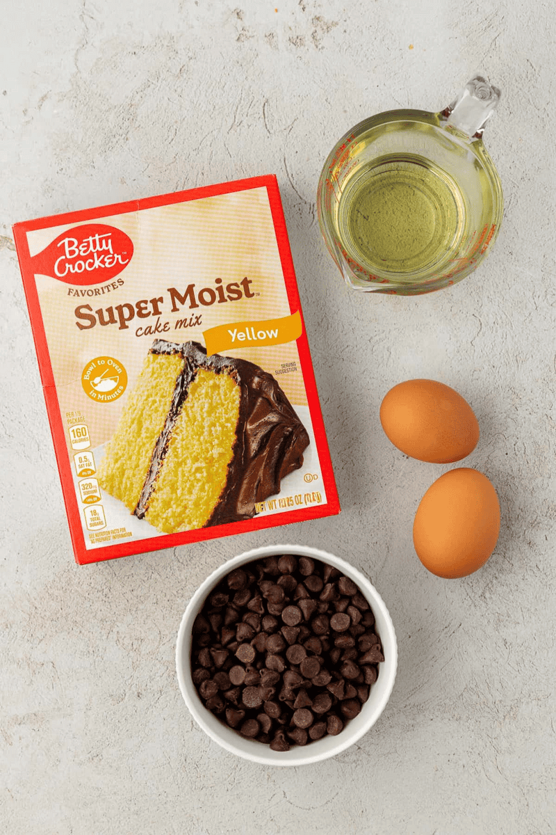 A box of Betty Crocker Super Moist yellow cake mix, a glass measuring cup with oil, two eggs, and a white bowl of chocolate chips—everything you need to make delicious cake mix cookies on a light countertop.