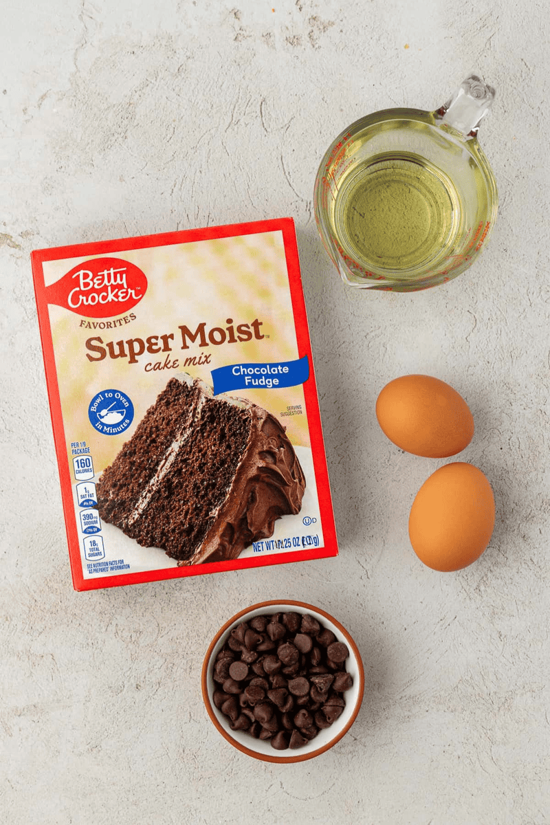 A box of Betty Crocker Super Moist Chocolate Fudge cake mix, two eggs, a cup of vegetable oil, and a small bowl of chocolate chips on a light textured surface—perfect for making delicious cake mix cookies.