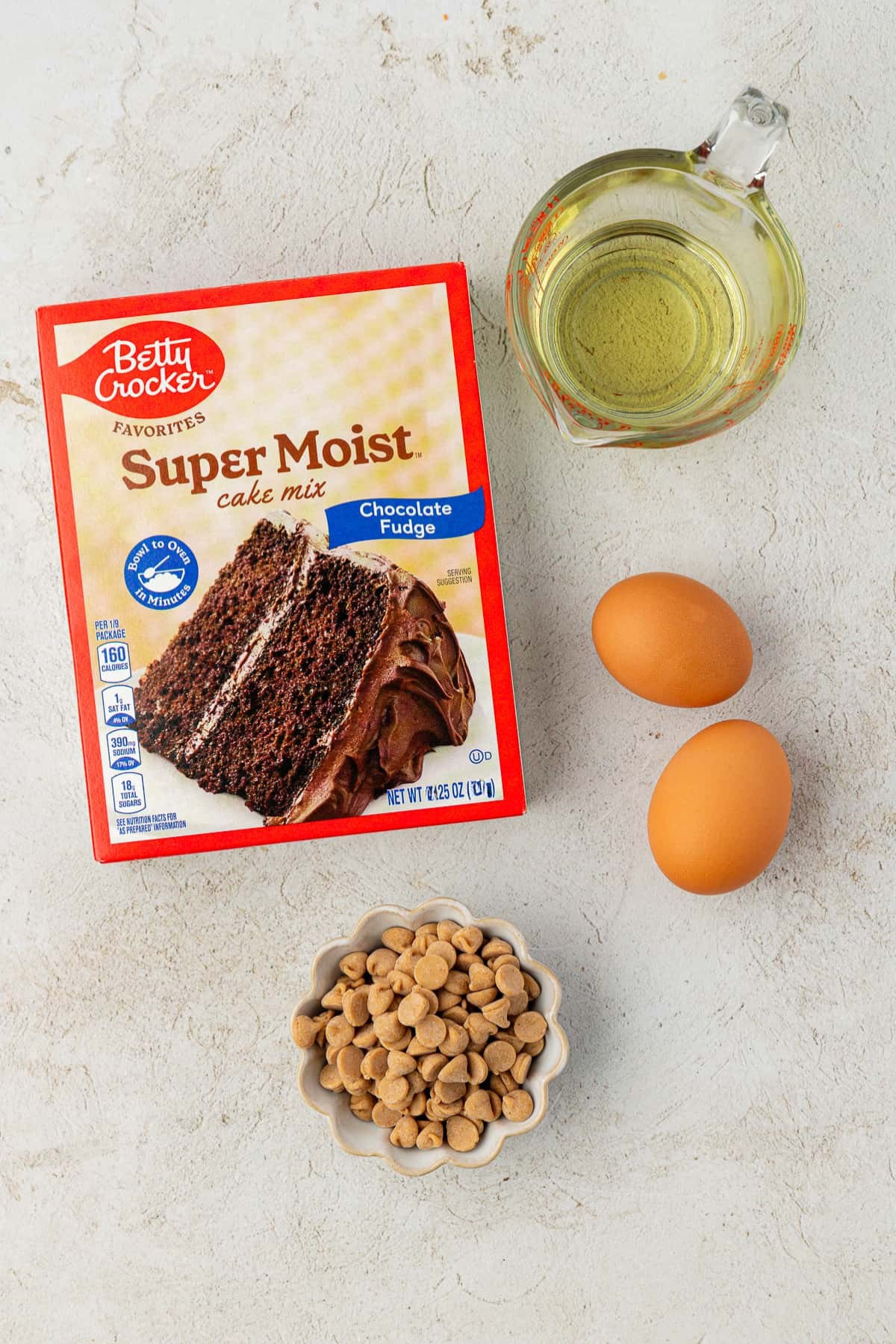 A box of Betty Crocker Super Moist chocolate fudge cake mix, a glass measuring cup with oil, two brown eggs, and a small bowl of peanut butter chips are arranged on a light countertop, ready to make delicious cake mix cookies.