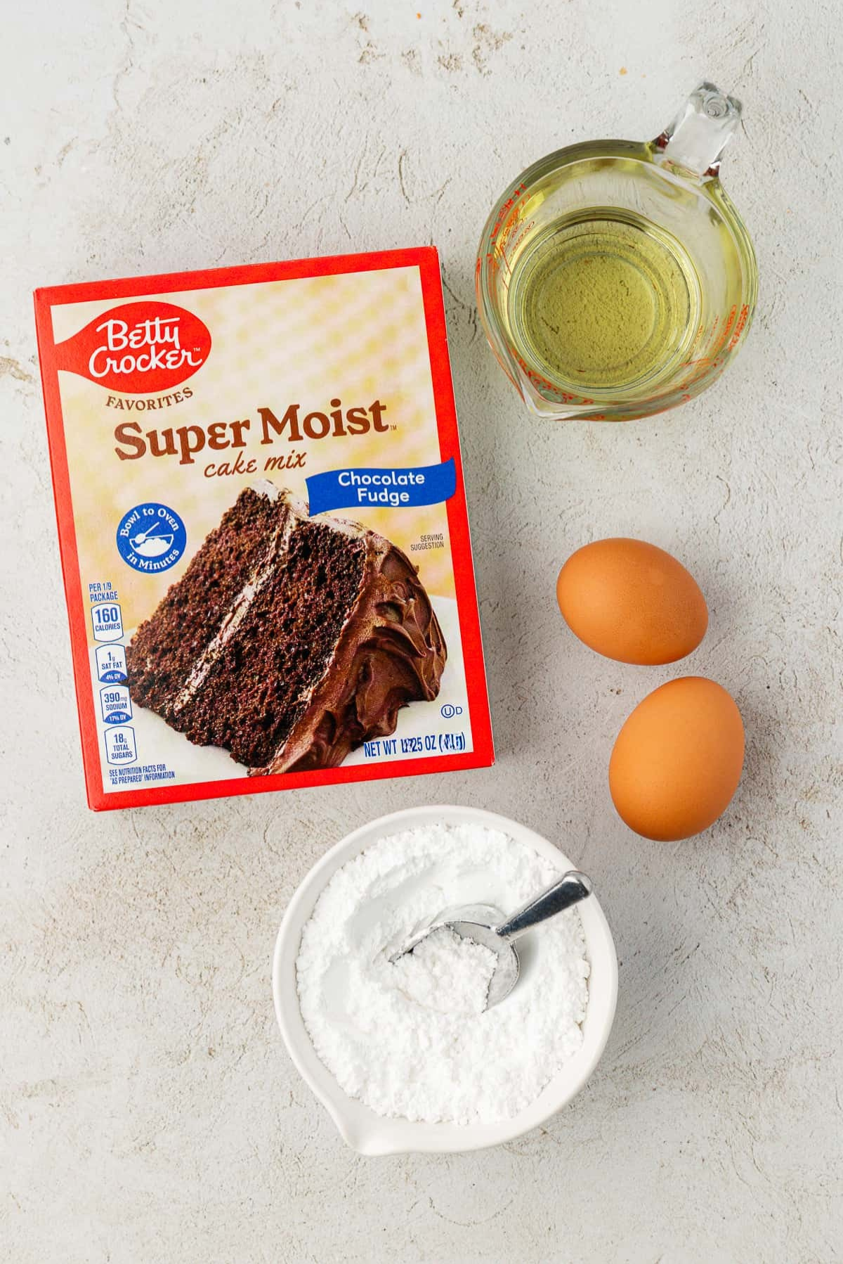A box of Betty Crocker Super Moist chocolate fudge cake mix, a glass measuring cup with oil, two eggs, and a bowl of powdered sugar with a spoon—everything you need to make delicious cake mix cookies on a light surface.
