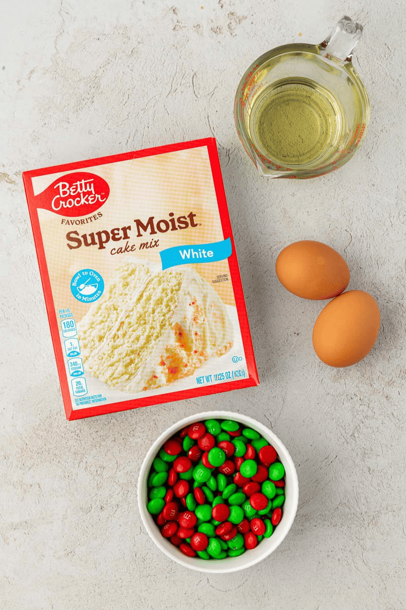 A box of Betty Crocker Super Moist white cake mix, two brown eggs, a glass measuring cup with oil, and a bowl of red and green M&M candies are arranged on a light surface—perfect for making festive cake mix cookies.